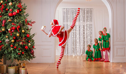 Children dressed as elves watch a dancing santa claus ballerina near a Christmas tree in a spacious white studio. - obrazy, fototapety, plakaty