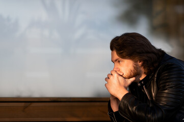 Pensive serious young bearded man wear black leather jacket sitting and thinking outdoors