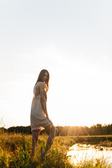 Obraz premium a young beautiful girl, of model appearance, in a shirt, stands among the landscape on the river bank, the setting sun illuminates her figure