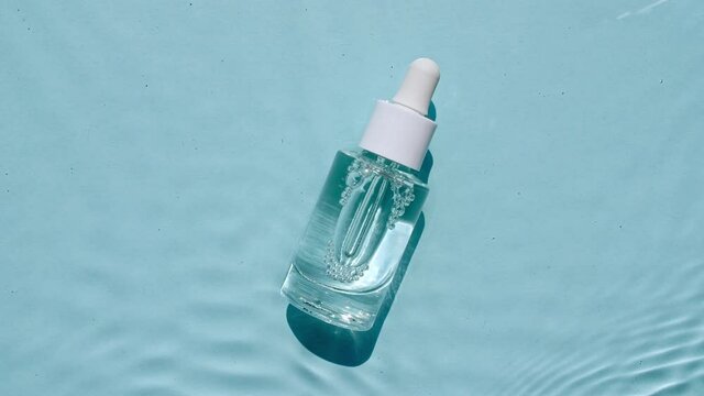 Glass cosmetic bottle with pipette lies on the surface of the water. Top view of water splash blue colored. Pure blue water with reflections sunlight. Sample of packaging for design, advertising.4k.