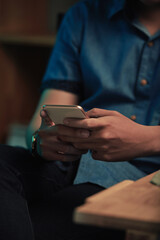 Close-up of unrecognizable man in casual shirt sitting at table and texting message on smartphone in dark room
