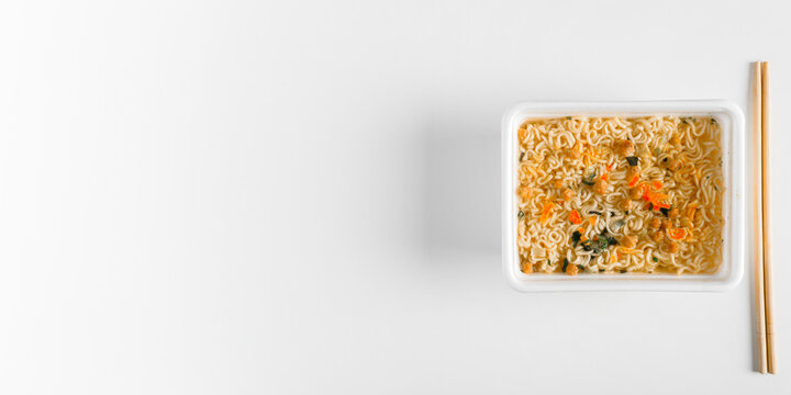 Chinese instant noodles in plastic box with wooden chopsticks on white table, banner with copy space for text. Top view ready-made azite dish with spices, unhealthy food