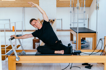 Healthy smiling blond man wearing sportswear practicing pilates in exercise studio on reformer special equipment. Caucasian male doing mermaid exercises on pilates reformer machines indoors.