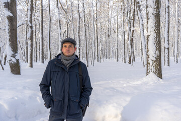 Fototapeta na wymiar Portrait of handsome middle-aged man walking in winter snowy park or forest. Attractive man in jacket, scarf and cap looking at camera. Winter mood, authentic lifestyle concept, stylish male outfit