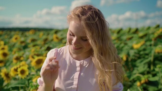 young beautiful pretty blonde caucasian girl in a long light dress in a sunflower field on a sunny day shyly smiles, straightens her curly hair