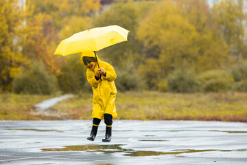 Boy in yellow waterproof cloak and black rubber boots with umbrella jump in a puddlein in park in...