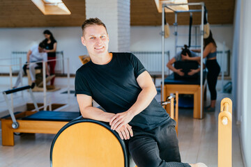 A confident pilates instructor and a healthy lifestyle. A handsome man does sports. The trainer portrait in pilates studio leaning on barrel.