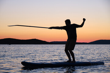 Silhouette of man on stand up paddleboard at sunset, pointing the paddle in a fencing position....