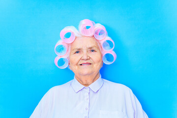 cute 80 years old woman with curlers on white hair looking happy in blue studio background