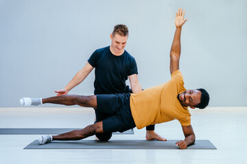 Male pilates trainer helps to african man, individual training participant, do right perform correctly. Extended different pose, class in sport club studio led by experienced man instructor concept.