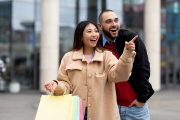 Portrait of smiling international couple holding shopper bags, pointing at store window, walking near shopping center