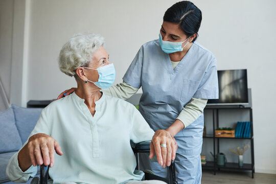 Portrait of female nurse assisting senior woman in wheelchair at home or in retirement center, both wearing masks