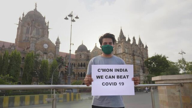 A young man wearing face mask standing and holding a placard with message 'C'mon India we can beat Covid 19' during city lockdown amid coronavirus corona against historic CSTM or VT terminus station