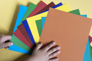 Children's hands hold sheets of colored paper on a colored background. The concept of children's creativity is handmade