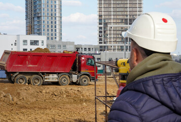 Photo of a construction worker in a white helmet who looks into the level at the construction site of multi-storey buildings with a red truck on the street in the city