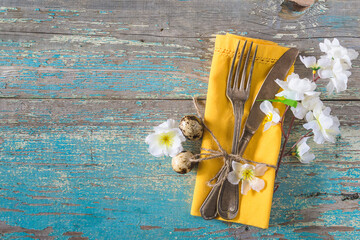 Festive place setting for easter holiday dinner: Vintage fork and knife on yellow napkin, white apple blossom on old blue paint wooden background, space for text - Powered by Adobe