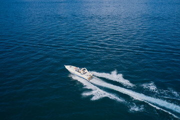 Large open white yacht with people moving fast diagonally on dark blue water aerial view.
