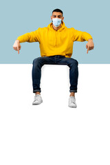 Young arab man in protective mask pointing at huge empty board for advertisement, sitting on it over blue background