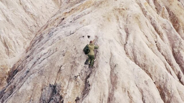 Guy with backpack climbs mountain slope made of clay, sand. Top, aerial view of man going up hill, moving along barkhan dune. Desert territory without plants, post-apocalypse, last person
