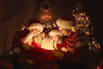 Fototapeta na wymiar Christmas Family opening Santa Bag with Gifts. Happy Parents and Kids sitting in front of Decorated Xmas Fir Tree and looking at Magic Light. Winter Holiday Eve