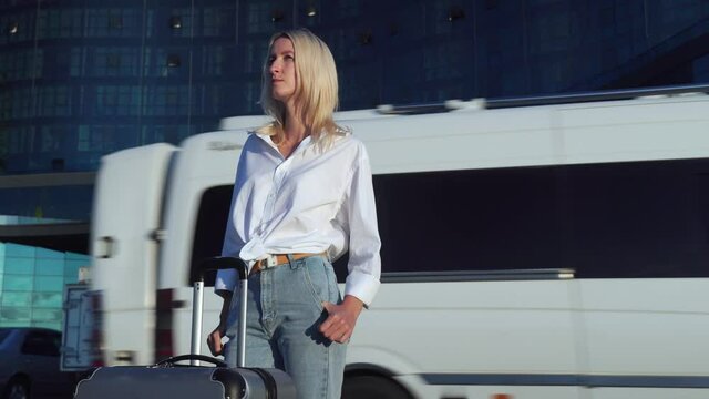girl with suitcase stands against background of city, tall skyscrapers and glass buildings. in background is parking lot, she is waiting for transfer or check-in at hotel. lady with luggage. of travel