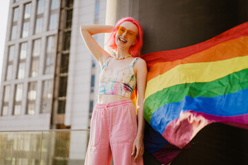 Young white woman laughing while standing with rainbow flag by wall