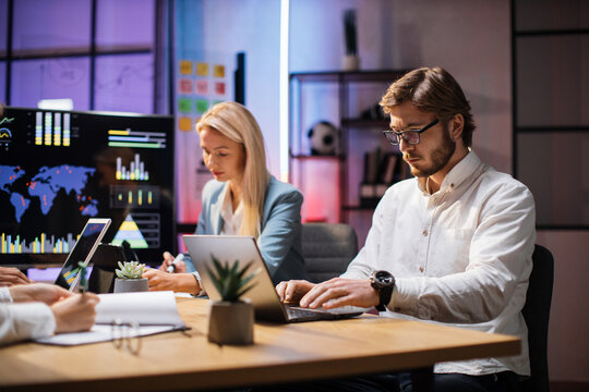 Focused caucasian man and woman in formal wear spending evening time at office for monitoring financial market situation. Qualified economists sitting at desk with modern laptop and tablet.