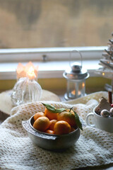 Silver bowl filled with fresh tangerines, various Christmas decorations, lit candles and soft blanket. Hygge at home. Selective focus.