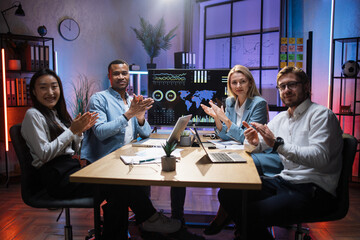 Multi ethnic male and female partners in formal clothes applauding together while sitting at office. Competent financiers with modern gadgets on table looking and smiling on camera.