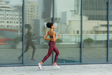 Fototapeta na wymiar Young biracial sportswoman in stylish tracksuit runs past modern building with glass facade reflecting large city street side view