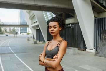 Concentrated biracial lady in stylish top with crossed arms stands against contemporary city building fence