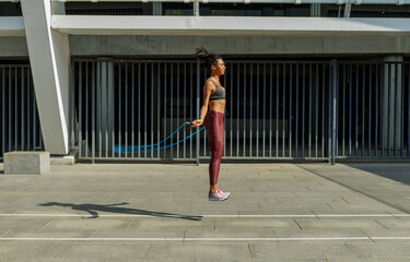 Fototapeta na wymiar Concentrated young woman in stylish tracksuit jumps rope near modern building on sunny city street
