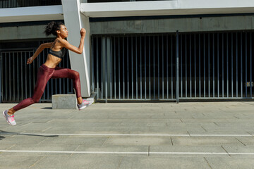 Strong biracial sportswoman in comfortable tracksuit runs jumping up past modern building on sunny city street side view