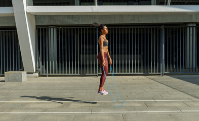Fototapeta na wymiar Concentrated young woman in stylish tracksuit jumps rope near modern building on sunny city street