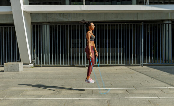 Concentrated young  woman in stylish tracksuit jumps rope near modern building on sunny city street - Powered by Adobe