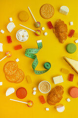Many different yummy candies with body measuring tape on yellow background, top view.