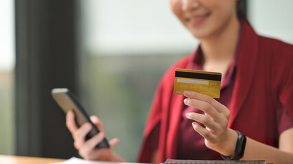 Photo of a young woman holding a credit card and smartphone in hand at the wooden working desk. Paying by credit card concept.