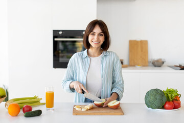 Happy young lady cutting fruits on wooden board, preparing fresh salad, standing in kitchen and...