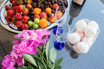 Summer picnic on the terrace with strawberries, cherries, peaches, marshmallows, peonies and a bottle of champagne and two glasses. Beautiful decor.