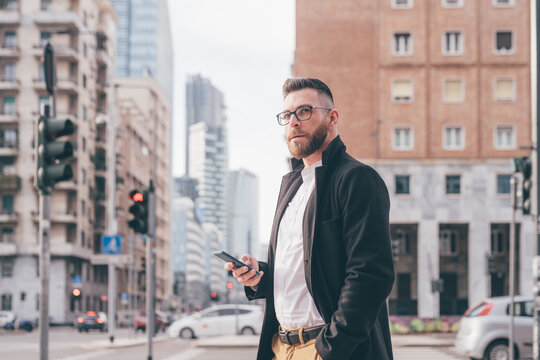 Young contemporary bearded businessman outdoor using smartphone overlooking confident and serene