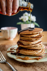 Hand pouring maple syrup on a healthy buckwheat pancake stack topped with chocolate and halvah on a decorative plate. Easy to make gluten free morning breakfast, brunch. Delicious golden pancake heap.