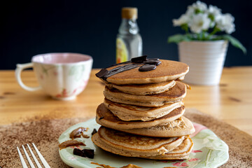 Healthy buckwheat pancake stack with dark chocolate, maple syrup and halvah on a decorative plate. Easy to make gluten free morning breakfast or brunch. Delicious heap of golden pancakes with teacup.