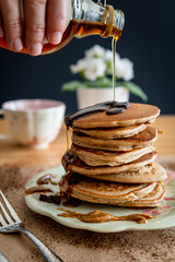 Hand pouring maple syrup on a healthy buckwheat pancake stack topped with chocolate and halvah on a decorative plate. Easy to make gluten free morning breakfast, brunch. Delicious golden pancake heap.