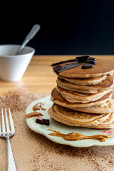 Healthy buckwheat pancake stack with chocolate, maple syrup and halvah on a decorative plate. Easy to make gluten free morning breakfast or brunch. Delicious heap of golden pancakes. dark background.