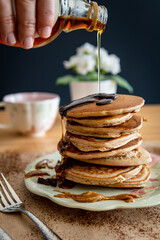 Hand pouring maple syrup on a healthy buckwheat pancake stack topped with chocolate and halvah on a...