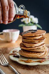Hand pouring maple syrup on a healthy buckwheat pancake stack topped with chocolate and halvah on a decorative plate. Easy to make gluten free morning breakfast, brunch. Delicious golden pancake heap.
