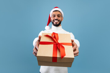 New Year celebration. Happy arab guy in Santa hat holding Christmas gift and stretching it to camera