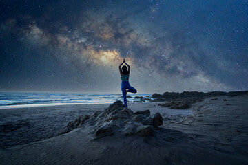 silhouette of a woman standing in yoga tree position on the rock or mountain with Milky Way...