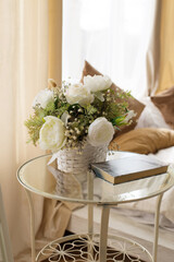 
White artificial roses in a basket on the coffee table, next to a book for reading. Interior decoration in pastel colors