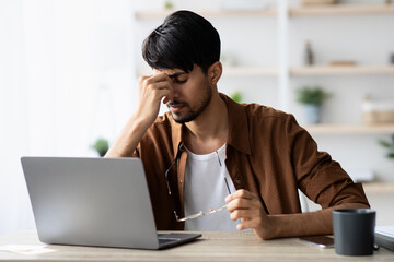 Tired indian man sitting in front of laptop, touching head